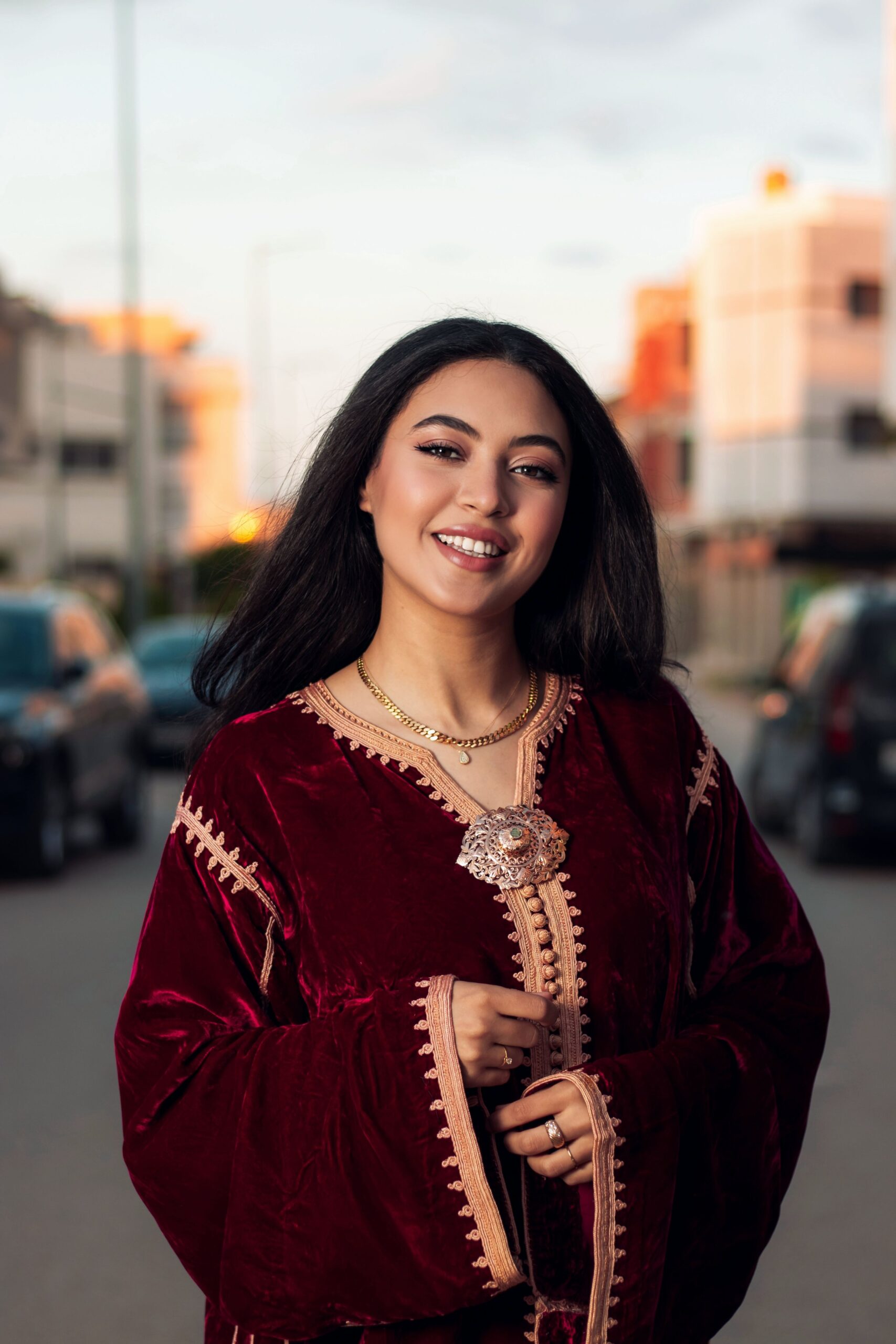 Elegant woman in a red velvet dress smiling on a street during sunset.